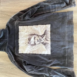 Svart velour hoodie Limitato  - Svart hoodie i mjuk velour med dragkedja och huva. På ryggen finns en stor beige patch med konstnärligt tryck av ett ansikte. Märke Limitato. 