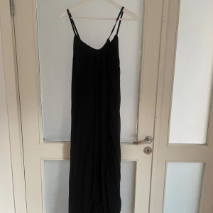 Svart långklänning från lager  157 - Svart långklänning från lager 157 med tunna axelband och avslappnad passform. Klänningen har en enkel och stilren design med lätt rynkad överdel och fallande tyg. Perfekt för dig som gillar minimalistisk stil och vill ha något bekvämt och snyggt.