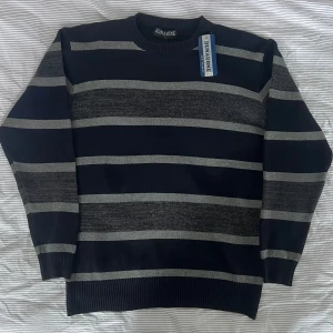 Randig tröja från Dunaone - Oanvänd och extremt snygg mörkblå cardigan från Dunaone med horisontella grå ränder. Tröjan har rund halsringning, ribbade muddar och är långärmad. Perfekt för dig som gillar klassisk stil med en modern twist.