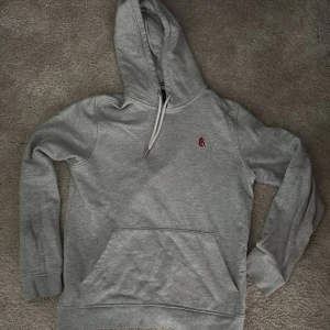 Grå hoodie från Bondelid  - Säljer en stilren grå hoodie från Bondelid med röd broderad logga på bröstet. Tröjan har huva med vit snörning och en stor magficka. Perfekt för dig som gillar klassisk och enkel streetstyle.