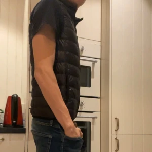 Svart dunväst från Ea7 Armani - Svart dunväst från Ea7 Armani med quiltad design och dragkedja framtill. Västen har en stilren look och passar perfekt över en hoodie eller t-shirt. Liten logga på bröstet och smidig passform som ger en sportig vibe.