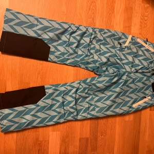 Mönstrade täckbyxor från Cross Sportswear - Snygga täckbyxor från Cross Sportswear i blått och ljusblått med grafiskt mönster. Byxorna har svarta förstärkningar vid bensluten, dragkedjor vid fickorna och är tillverkade i slitstarkt syntetmaterial. Perfekta för vinterns alla äventyr.