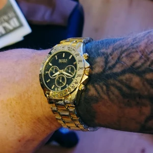 HUGO BOSS Guldklocka Helt ny med skyddsplast kvar  - Helt ny kronografklocka från Hugo Boss med guldfärgad länk och svart urtavla. Fortfarande förseglad i plast endast provat på för bilder. Klockan har tre mindre urtavlor, tydliga index och detaljerade knappar på sidan. Stilen är sportig men ändå lyxig, perfekt för dig som gillar statement-accessoarer.