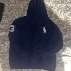 Blå hoodie från Polo Ralph Lauren - Säljer en blå hoodie från Polo Ralph Lauren med stor vit logga på bröstet och siffra 3 på ärmen. Klassisk känguruficka framtill och huva. Perfekt för dig som gillar sportig och stilren streetwear.storlek L men passar M är 194 och är lite över midjan 