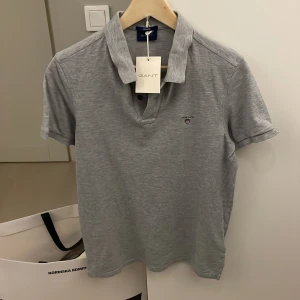 Grå pikétröja från GANT - Snygg grå pikétröja från GANT med klassisk krage och korta ärmar. Tröjan har diskret logga broderad på bröstet och är tillverkad i mjuk bomull. Perfekt för en clean och stilren look.