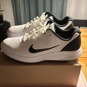 Vita och svarta Nike sneakers - Snygga vita golf skor från Nike med svart swoosh och svarta detaljer på häl och sula. Skorna har snörning, rund tå och är tillverkade i syntetmaterial med textilfoder. Perfekta för dig som gillar stilrena och sportiga skor.