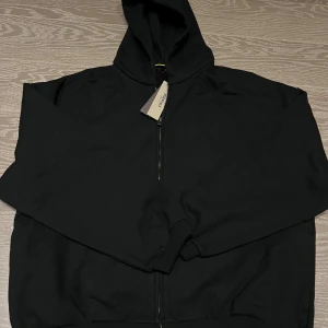 Svart hoodie från Fear of God Essentials - Svart hoodie från Fear of God Essentials med dragkedja och huva. Stora vita bokstäver 'FEAR OF GOD STATE' på ryggen. Tillverkad i 100% bomull och har en clean, oversized vibe. Perfekt för dig som gillar streetwear och vill sticka ut.