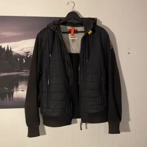 Svart pufferjacka från Parajumpers M - Svart cardigan från Parajumpers i storlek M. Jackan har quiltad front, mjuka fleeceärmar och huva. Snygga detaljer som orange hängare och dragkedja framtill. Perfekt för dig som vill ha snygg cardigan nu på hösten🍂🍁
