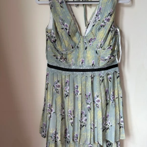 Blommig långklänning från Self-Portrait - Säljer en romantisk långklänning från Self-Portrait med blommigt mönster i lila och svart på ljusgrön botten. Klänningen har spetsdetaljer, v-ringning och omlottband i ryggen. Tillverkad i lätt chiffong med lager på lager-effekt och markerad midja med spets.