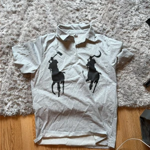 Grå pikétröja med svart tryck Storlek L - Säljer en ljusgrå pikétröja i bomull strlk L med två stora svarta polospelare tryckta på framsidan. Klassisk krage och korta ärmar, perfekt för en chill och stilren look. Passar dig som gillar sportiga och trendiga vibes. Aldrig använd