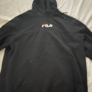 Svart hoodie från FILA med logga - Svart hoodie från FILA med broderad logga i vitt och rött på bröstet. Klassisk känguruficka framtill och justerbar huva med snörning. Perfekt för chill dagar eller streetwear-stil.