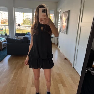 Svart omlottklänning med volang - Säljer en svart kort omlottklänning med volangkant och öppen rygg. Klänningen har en hög hals med knyt och är ärmlös, vilket ger en cool och trendig look. Materialet är lätt och luftigt, perfekt för varmare dagar eller när du vill sticka ut.