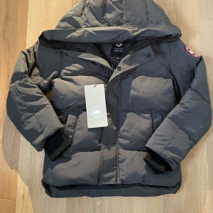 Mörkgrå dunjacka från Canada Goose XS - Snygg mörkgrå dunjacka från Canada Goose i storlek XS. Jackan har huva, dragkedja och knappar framtill, samt två stora fickor. Klassisk patch på ärmen och ribbade muddar vid ärmslut. Perfekt för kalla vinterdagar.