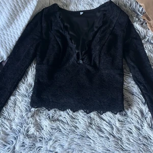 Svart spetsblus med v-ringning - Säljer en svart blus i spets med lång ärm och v-ringad hals. Toppen har fina spetsdetaljer över hela och en lätt croppad passform. Perfekt för dig som gillar en stilren men ändå edgy look.