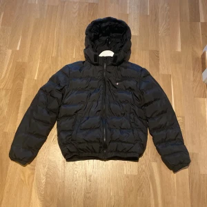 Svart pufferjacka från Gant med huva - Svart pufferjacka från Gant med huva och dragkedja framtill. Jackan är vadderad och har en klassisk, sportig look med elastiska muddar vid ärmslut och nederkant. Perfekt för kalla dagar och enkel att matcha med allt. Men det finns en defekt, märket där det står Gant på  sitter löst på ena sidan. Men annars är det ingenting fel på varan👍