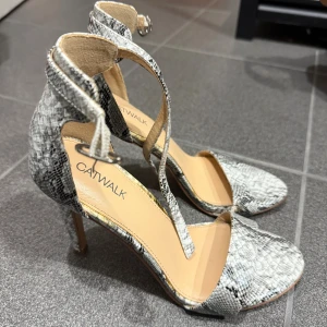 Catwalk sandaler med ormskinnsmönster - Snygga sandaler från Catwalk med höga klackar och trendigt ormskinnsmönster i svart, grått och vitt. Skorna har öppen tå, smal rem över foten och justerbar ankelrem med spänne. Perfekta för dig som vill sticka ut med en cool och modern look. pris går att diskutera 