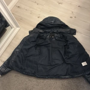 Svart pufferjacka från 157, stl S - Snygg marinblå pufferjacka från 157 med avtagbar huva och dragkedja framtill. Jackan har quiltad design, två fickor med dragkedja och en glansig finish. Perfekt för kyliga dagar och enkel att matcha med olika outfits.