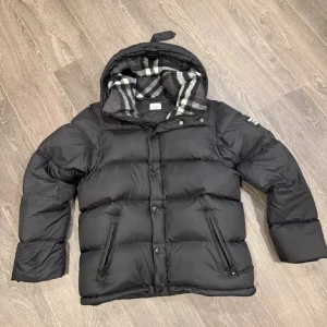 Svart dunjacka från Burberry - Svart pufferjacka från Burberry med stor logopatch på ärmen och klassiskt rutigt foder i huvan. Jackan har dragkedja och knappar framtill, två fickor med dragkedja och en riktigt fluffig look. Perfekt för kalla dagar när du vill vara både varm och snygg.