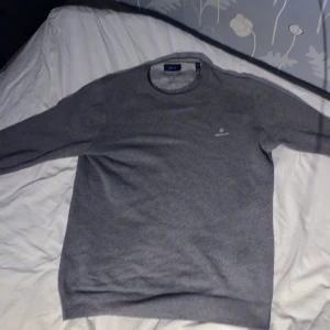 Grå stickad tröja från Gant - Säljer en stilren grå stickad tröja från Gant med rund halsringning och diskret logga på bröstet. Perfekt för lager-på-lager och passar till många olika outfits. Tröjan har långa ärmar och är mjuk och bekväm.