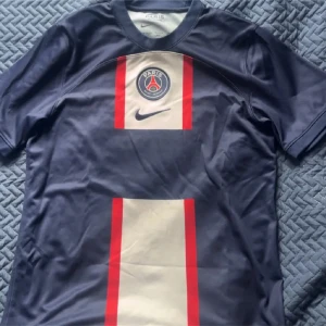 PSG Nike fotbollströja marinblå M - Paris Saint-Germain fotbollströja från Nike i marinblått med breda vertikala ränder i vitt och röda kanter. Tröjan har PSG-logga och Nike Swoosh på bröstet samt är tillverkad i Dri-FIT-material som andas. Klassisk rund hals och korta ärmar.