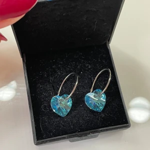 Blå hjärtformade örhängen i silver - Supersöta örhängen med gnistrande blå hjärtan i kristall-look och tunna silverfärgade ringar. Perfekta för dig som vill ha något unikt och glittrigt som sticker ut. Hjärtformade hängen ger en romantisk vibe och passar till alla tillfällen.