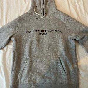 Grå hoodie från Tommy Hilfiger - Säljer en grå hoodie från Tommy Hilfiger med klassisk logga och text på bröstet. Tröjan har huva med snörning och en stor magficka. Perfekt för dig som gillar en clean och sportig stil.