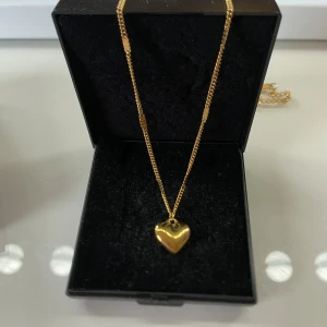 Guldigt halsband med hjärthänge - Supersött halsband i guldton med ett glänsande hjärthänge. Kedjan har små dekorativa detaljer längs hela längden och ett klassiskt lås. Perfekt accessoar för att lyfta vilken outfit som helst med en romantisk touch.