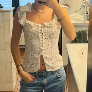 Vit blommig blus med volang - Säljer en söt vit blus med blommigt mönster och volangdetaljer vid axlar och halsringning. Blusen har knappar framtill och är croppad med en lätt v-ringad form. Perfekt för dig som gillar romantisk och feminin stil.
