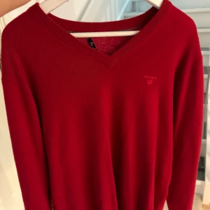 Röd v-ringad tröja från GANT - Röd v-ringad tröja från GANT med diskret broderad logga på bröstet. Tröjan har långa ärmar och är tillverkad i mjuk bomull, perfekt för lager-på-lager. Klassisk och stilren modell som funkar till allt.