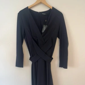 Mörkblå omlottklänning Tiger of Sweden - Snygg mörkblå klänning från Tiger of Sweden med omlottdesign och trekvartsärm. Klänningen har v-ringning och markerad midja med korsade detaljer framtill. Stängs med dragkedja bak. Perfekt för dig som gillar stilrena och eleganta plagg. Prislapp kvar, nypris 999kr