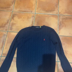 Mörkblå kabelstickad tröja Polo Ralph Lauren - Kabelstickad mörkblå tröja från Polo Ralph Lauren med klassisk rund halsringning och röd broderad logga på bröstet. Långärmad och i mjuk bomull, perfekt för lager-på-lager under höst och vinter.