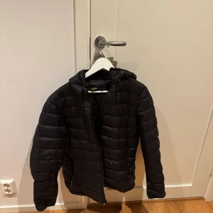 Svart Ralph lauren pufferjacka med huva - Svart pufferjacka med huva och dragkedja framtill. Jackan har horisontella sömmar, två fickor med dragkedja och en klassisk, enkel design. Perfekt för kyliga dagar och enkel att matcha med olika outfits.