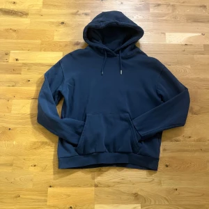 Mörkblå hoodie från RYVLS - Mörkblå hoodie från RYVLS med klassisk känguruficka och dragsko i huvan. Tröjan har en relaxed passform och är tillverkad i mjuk bomullsblandning, perfekt för kyliga dagar. Enkel och stilren design som funkar till det mesta.