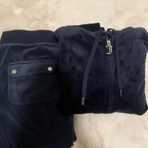 Mörkblå velour hoodie från Juicy Couture - Mysig mörkblå hoodie i mjuk velour från Juicy Couture med dragkedja, snörning i huvan och ikoniskt JC-hänge. Klassisk passform och perfekt för chill dagar. Snygga detaljer och broderad logga på bröstet. En metal grej på snöret har lossnat annars bra skick, lite använt. Tröjan i storlek S och byxorna i XS