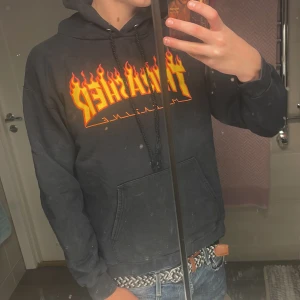 Svart Thrasher Flame hoodie - Svart hoodie från Thrasher med ikoniskt Flame-logga i gult och rött framtill. Klassisk känguruficka, huva med snörning och mjukt material. Perfekt för dig som gillar streetwear och skatekänsla.
