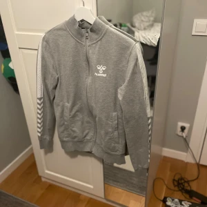 Grå ziptröja från Hummel S - Grå ziptröja från Hummel med vita detaljer och logga på bröstet. Tröjan har hel dragkedja, ribbade muddar och klassiska Hummel-pilar längs ärmarna. Perfekt för en sportig look eller chill vardagsstil.