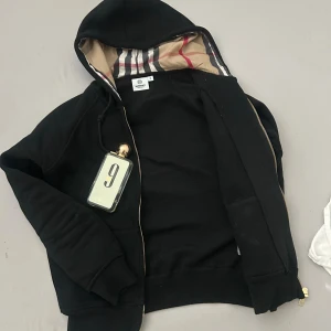 Svart hoodiejacka från Burberry - Svart hoodiejacka från Burberry med dragkedja och klassiskt rutigt foder i huvan. Jackan har ribbade muddar, snörning i huvan och är tillverkad i mjuk bomull. Perfekt för dig som gillar stilrena och exklusiva streetwear-plagg.