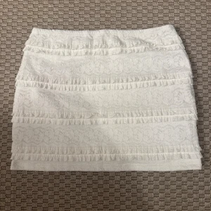 Vit spetskjol med volangdetaljer - Aldrig använt, köpt i Spanien. Supersöt vit kortkjol med spets och flera volangrader runt om. Kjolen har en feminin och romantisk känsla med sitt mönstrade tyg och de små volangerna som ger extra liv. Perfekt för dig som gillar en ljus och fräsch stil.