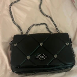 Svart quiltad axelväska Love Moschino - Snygg svart quiltad axelväska från Love Moschino med hjärtformade metalldetaljer och kedjerem. Väskan har en stilren design och logga framtill, perfekt för dig som vill ha en trendig accessoar med det lilla extra.