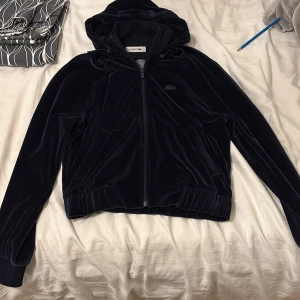 Mörkblå velourjacka från Lacoste - Mörkblå velourjacka från Lacoste med huva och dragkedja framtill. Klassisk krokodillogga på bröstet, ribbade muddar och två fickor. Jackan har en mjuk och lyxig känsla, perfekt för dig som gillar sportig och stilren look.
