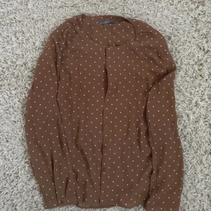 Brun prickig kofta med knappar - Säljer en brun kofta med vita prickar och knäppning framtill. Koftan har lång ärm och en klassisk rund halsringning. Perfekt för dig som gillar retrovibbar och vill ha något som sticker ut lite extra i garderoben🤩