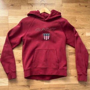 Röd hoodie från GANT med brodyr - Snygg röd hoodie från GANT med broderad logga och sköld på bröstet. Tröjan har huva, känguruficka och långa ärmar. Tillverkad i mjuk bomullsmix, perfekt för chill dagar eller när du vill ha en sportig look. Storlek M men passar S, skriv om ni har några frågor.