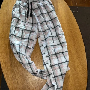 Rutiga mjukisbyxor från CRW, S - Säljer ett par superbekväma mjukisbyxor från CRW i storlek S. Byxorna är vita med rosa och grått rutmönster, har resår i midjan och svarta snören för justering. Perfekta för chill dagar hemma eller när du vill vara extra comfy.