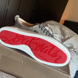 Grå mockasneakers från Louboutin - Snygga grå sneakers från Christian Louboutin i mjuk mocka med klassisk röd sula och vita kanter. Skorna har rund tå, platt sula och breda grå snören. Perfekta för dig som vill sticka ut med exklusiv streetstyle. Priset säger mycket. 