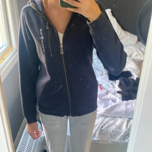 Mörkblå hoodie från Sail Racing - Mörkblå hoodie från Sail Racing med huva och hel dragkedja framtill. Tröjan har en snygg logga på bröstet och en extra dragkedjeficka. Mjuk insida och ribbade muddar vid ärmslut och nederkant. Perfekt för chill eller när du vill vara lite sportig.