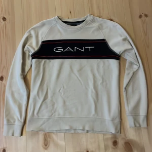 Vit GANT tröja med logga.  - Snygg beige sweatshirt från GANT med svart panel och vit logga över bröstet, samt röda linjer som detalj. Tröjan har rund halsringning och långa ärmar. Perfekt för en chill och stilren look.