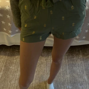 Gröna shorts - Säljer ett par gröna shorts med guldiga ananastryck från H&M, de har snörning med tofsar i midjan. De har en loose passform och elastisk midja med volangkant upptill. Kanske använda en gång så väldigt bra skick. Det står stl 146/152 men passar mig som har stl XS-S i vanliga fall. Hör gärna av er om frågor!❤️