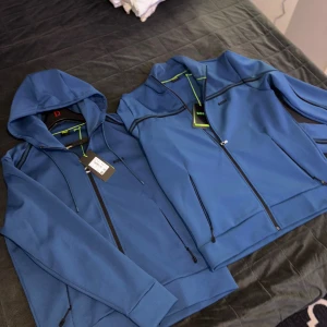 Blå zipjacka från BOSS med huva - 2st blåa ziphoodies från BOSS helt nya oanvända med svarta detaljer och dragkedjor. En jacka har huva med snörning, den andra har ståkrage. Båda har svarta linjer längs axlar och bröst, samt BOSS-logga på bröstet. Perfekt för en sportig och stilren look.