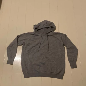Grå hoodie med snörning - En enkel grå stickad tröja med huva och snörning framtill. Tröjan har långärmade ribbade muddar och är tillverkad i mjukt och skönt stickat material. Perfekt för chill dagar och passar dig som gillar basic och stilrent. Storlek S men är mindre i storlek. Färgen ser mörk ut på bild men är ljusare i verkligheten