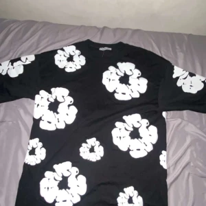 Svart t-shirt med vita blommor Mastiff - Svart t-shirt från Mastiff i storlek S med stora vita blommönster över hela plagget. T-shirten har rund hals och korta ärmar. Materialet är mjuk bomull som känns skönt mot huden. Perfekt för dig som gillar grafiska prints och vill sticka ut.